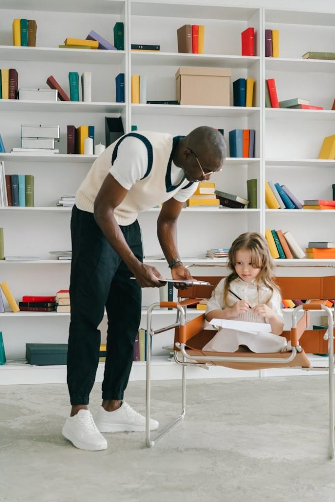 An adult male teacher assists a young girl in a classroom setting.